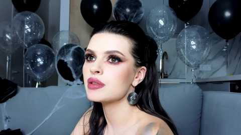 Video of a woman with fair skin, dark hair, wearing black earrings and bold red lipstick, surrounded by black and silver balloons.