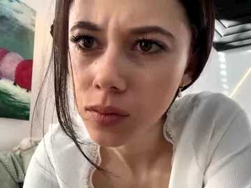 Video of a Caucasian woman with dark hair, wearing a white shirt, looking serious with a slight frown. Background includes a green abstract painting and a ceiling light.