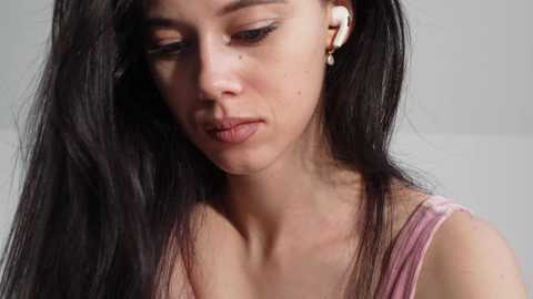 Video of a young woman with long black hair, fair skin, and a slender build, wearing a pink tank top and white earbuds, looking introspective against a plain, light-colored background.