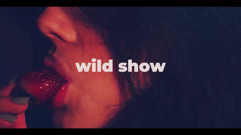 Video of a close-up of a person with dark hair, eating a strawberry with a red lipstick smear on their mouth. The background is dark, and the text \"wild show\" is overlaid in white.