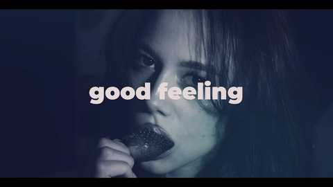 Video of a young woman with long, straight hair, half-lit in dark, moody lighting, holding a black object to her lips, with the text \"good feeling\" overlaid in white.