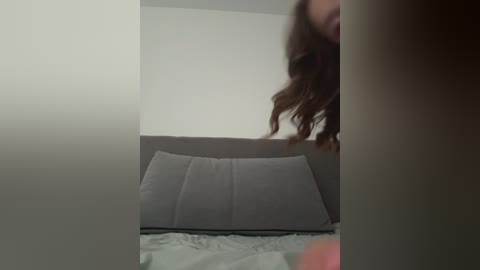 A video of a woman with long, wavy brown hair partially visible, standing in a dimly lit room with a grey bed and pillows. The background is plain and white.