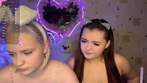 Video of two young women with light skin, wearing cat ears, in a cozy room with heart-shaped neon lights and a poster on the wall.