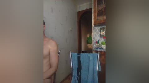 Video of a nude, muscular man standing in a dimly lit, unfinished bathroom with beige walls and a wooden door. A blue towel hangs on a rack.