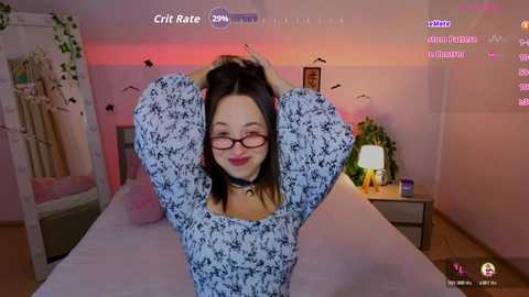 Video of a young woman with straight brown hair, wearing glasses, a floral dress, and a black choker, posing with her arms raised in a bedroom with pink walls, a bed, and a green plant.