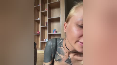 Video of a blonde woman with a black tattoo of geometric shapes on her chest, wearing a black choker, smiling, in a room with wooden shelves filled with small figurines.