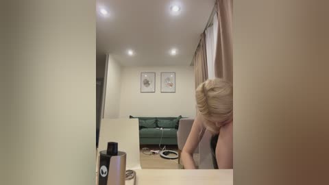 Video of a blonde woman in a beige room, seen from behind, leaning over a light-colored table with a black bottle. Green sofa, framed art on walls, beige curtains, and ceiling lights.