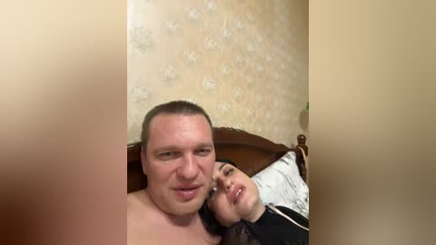 Video of a man and woman lying on a bed, man topless, woman partially covered, beige wallpaper, blurred background.