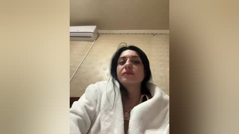 A video of a woman with long black hair, wearing a white robe, sitting on a beige couch in a dimly lit room with a white air conditioner on the wall.