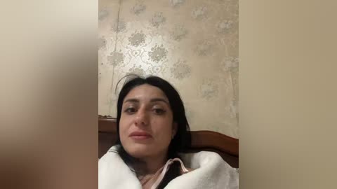 Video of a woman with long dark hair, lying on a bed with a beige floral wallpaper in the background. She is wrapped in a white blanket, her expression neutral.