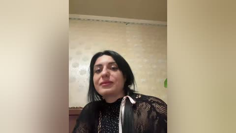 Video of a woman with long black hair, fair skin, and light makeup, wearing a black lace blouse and a black tie, standing in front of a beige, patterned wallpaper.