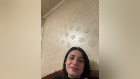 A video of a woman with long black hair, wearing a black top, sitting against a beige wall with a floral pattern. Her expression is neutral.