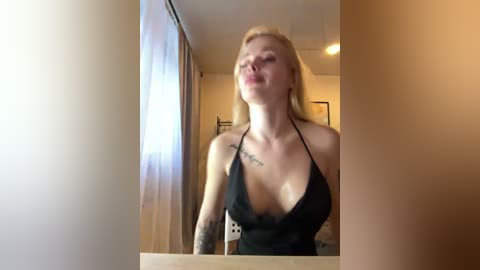Video of a blonde Caucasian woman in a low-cut black halter top, leaning against a bathroom mirror, with a tattoo on her left shoulder. Background features a window with white curtains and a beige wall.