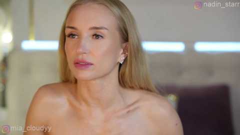 Video of a fair-skinned, blonde woman with long hair, wearing small diamond earrings, sitting topless, looking thoughtful. Background features a blurred modern room with a tufted headboard and a purple cushion.