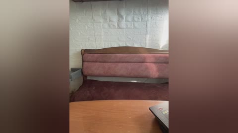 A video of a small, dimly lit room with a pink upholstered bed against a white textured wall, a wooden desk, and a TV remote on the desk.