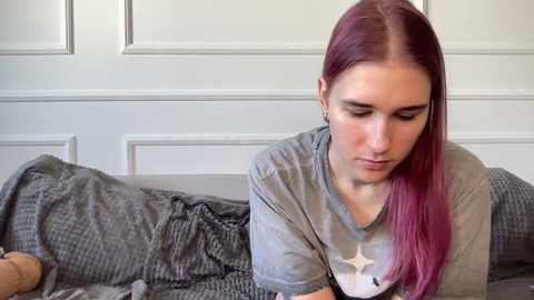 Video of a young woman with long, straight, pink hair, wearing a gray t-shirt, sitting on a gray, textured blanket on a couch, in a softly lit room with white paneled walls.