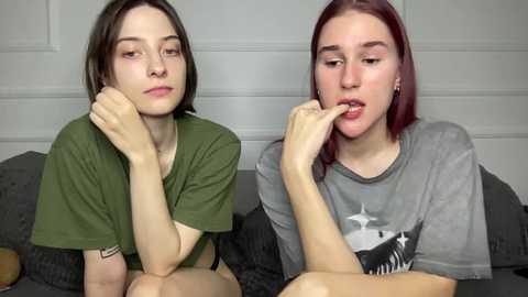 Video of two young women with pale skin, one with dark hair, the other with red hair, sitting on a bed, wearing casual t-shirts, in a simple, neutral bedroom setting.
