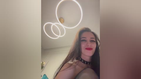 Video of a young woman with long, straight brown hair, wearing red lipstick and a black choker, standing in a modern, dimly lit room with circular white lights on the ceiling.