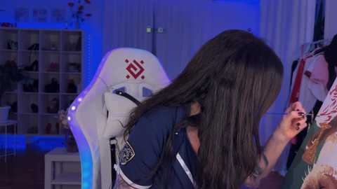 Video of a person with long black hair, wearing a dark shirt, sitting in a white gaming chair, with a tattoo on their arm, in a dimly lit room with blue lighting, shelves, and clothes.