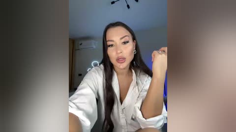 Video of a young woman with long dark hair, wearing a white robe, posing seductively in a dimly lit room.