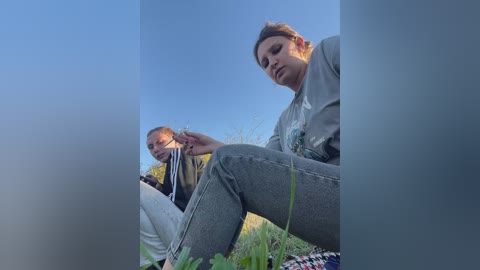 Video of a young woman in a beige jacket and a man in a yellow shirt, sitting in a grassy field with a picnic blanket, under a clear blue sky.