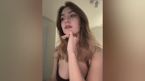 Video of a fair-skinned woman with long, light brown hair, wearing a dark top, with a thoughtful expression, indoors, against a beige wall, holding her chin.