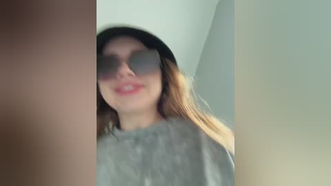 Video of a young woman with light skin, long brown hair, wearing dark sunglasses and a gray shirt. The image is slightly out of focus and features a blurred background.