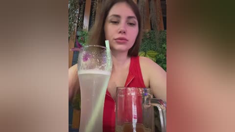 Video of a young woman with fair skin and long brown hair, wearing a red sleeveless top, sipping a tall glass of lemonade with a straw, seated outdoors amidst lush greenery and wooden structures.