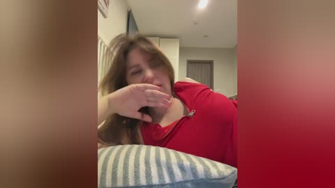 Video of a woman with long brown hair in a red shirt, leaning on a striped pillow, in a dimly lit room with beige walls and a closed door.