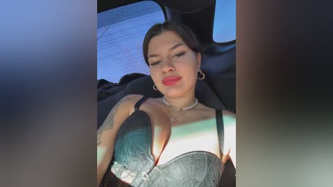Video of a young woman with medium skin tone, dark hair, and a tattoo on her left arm, wearing a black lace bra, sitting in a car.