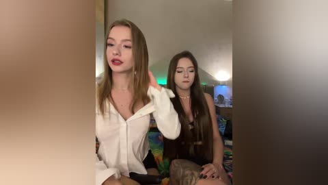 Video of two young women in a dimly-lit bedroom, one with long, straight brown hair and a white shirt, the other with long, wavy brown hair and a black top.