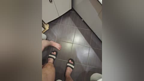 A video captures two barefoot individuals in a modern bathroom, with dark tiled floors and white cabinetry. The view is from a low angle, showcasing the feet and lower legs.