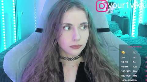 Video of a young woman with long, wavy brown hair, wearing a black choker and black top, seated in a dimly lit room with blue LED strip lights and a gray cushion behind her.