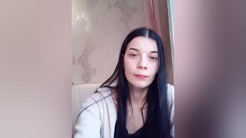 A video of a young woman with long, straight black hair, fair skin, and light makeup, wearing a white coat over a black top, standing in front of a white bathroom mirror.