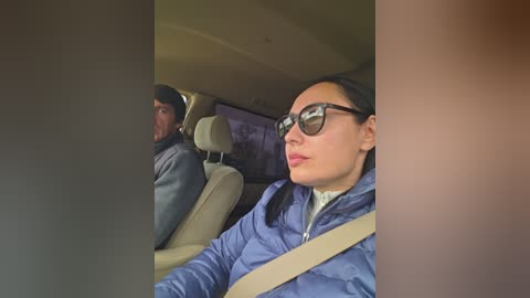 A video of an Asian woman with dark hair, wearing sunglasses and a blue puffer jacket, riding in a car with a man in the background.