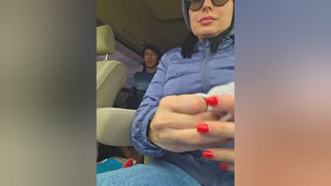 A video shows a woman with red nails and black hair, wearing glasses, a blue jacket, and a light sweater, sitting in a car next to a man with glasses.