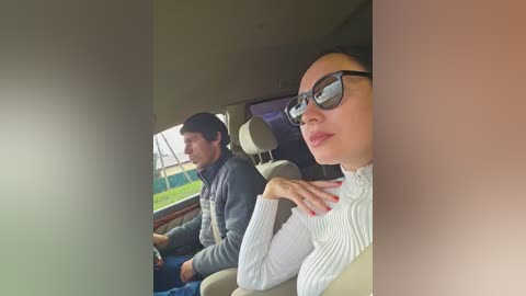 Video of a woman with dark hair, glasses, and a white sweater, sitting beside a man in a gray jacket in a car.