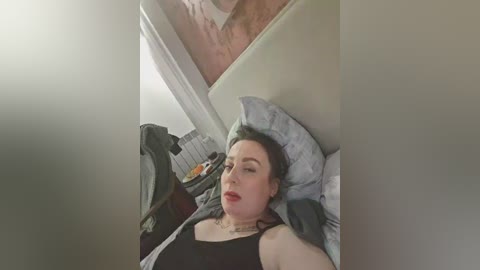 Video of a woman with light skin and dark hair lying in bed, wearing a black tank top, with a gray blanket and pillows.