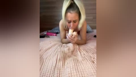 A video of a topless blonde woman with a medium build and small breasts, lying on a pink quilted bed, holding a banana in her mouth, with a wooden headboard in the background.