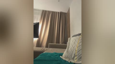 Video of a simple, cozy bedroom with beige curtains, a bed with a green blanket, and a yellow pillow, featuring a flat-screen TV mounted on the wall.