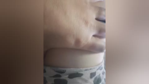 A video captures a close-up of a person's face and chest. The individual has light skin, visible breast, and wears a gray, polka-dotted top. The image is slightly blurry, with soft lighting.