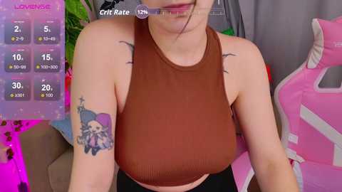 Video of a light-skinned woman with large breasts, wearing a brown ribbed tank top, featuring a colorful anime character tattoo on her upper arm. Background includes gaming gear, and virtual overlays.