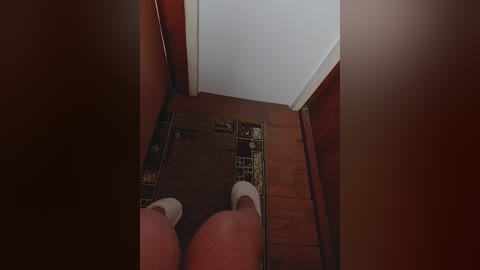 A video from a low angle shows a person's feet in white socks standing on a wooden floor, with a brown and black patterned rug. A doorway with red walls and white trim is visible.