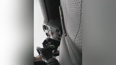 Video of a car interior showing a dashboard with multiple dials, gearshift, and pedals. The car seat is upholstered in light gray fabric with perforated detailing. A green keychain is visible on the gearshift.