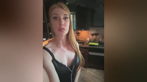 Video of a fair-skinned, blonde woman with long hair, wearing a black lace bra, in a dimly lit modern kitchen.