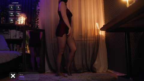A video of a slender, fair-skinned woman in a sheer black lingerie dress standing beside a dimly-lit window, with a digital interface displaying \"X\" and \"O\" in the bottom-left corner.
