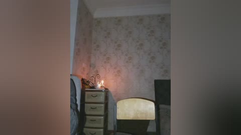 Video of a dimly lit, vintage bedroom with floral wallpaper, a beige dresser, a lit candle, a metal-framed chair, and a bed visible through a doorway.