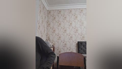 A video of a small, dimly lit room with beige floral wallpaper, a wooden chair, and a desk. The scene is viewed from the corner, with a blurry figure in a dark jacket partially visible.