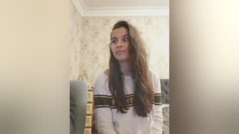 A video of a young woman with long, wavy brown hair, wearing a white shirt with black and gold stripes, sitting in a cozy, beige room with floral wallpaper.