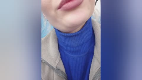 A close-up video of a person's chin, lips, and neck, wearing a blue ribbed sweater under a beige zip-up jacket, with a blurred background.